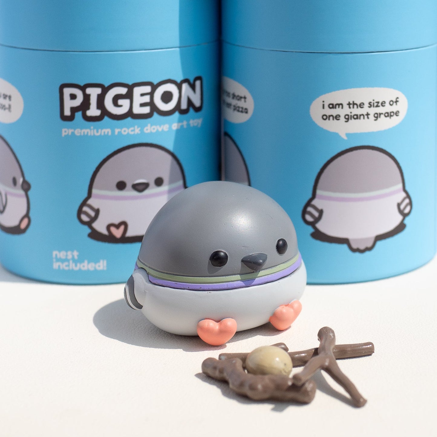 A small cartoon pigeon art toy by pigeoncrafts, in front of two blue cylindrical packaging containers labeled 'PIGEON' and 'nest included!'. 