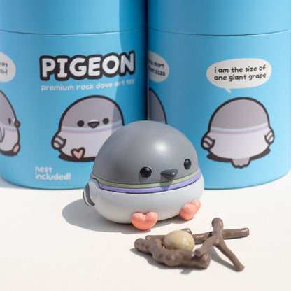 A small cartoon pigeon art toy by pigeoncrafts, in front of two blue cylindrical packaging containers labeled 'PIGEON' and 'nest included!'. 
